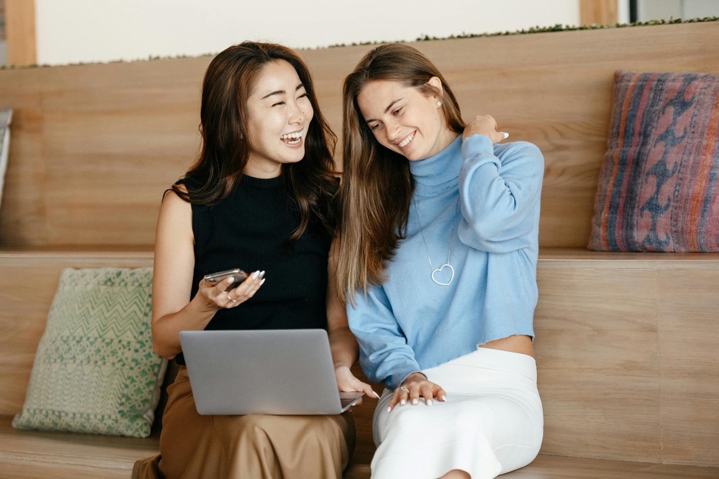 Two women sitting on a couch, laughing while looking at a laptop screen. One of them has a cell phone in her right hand. They’re using their home WiFi connection to access the internet.