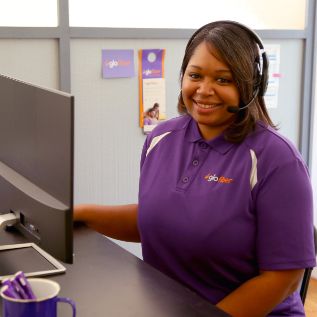 A smiling Glo Fiber Customer Service Representative wearing a headset and sitting at a desk with a computer